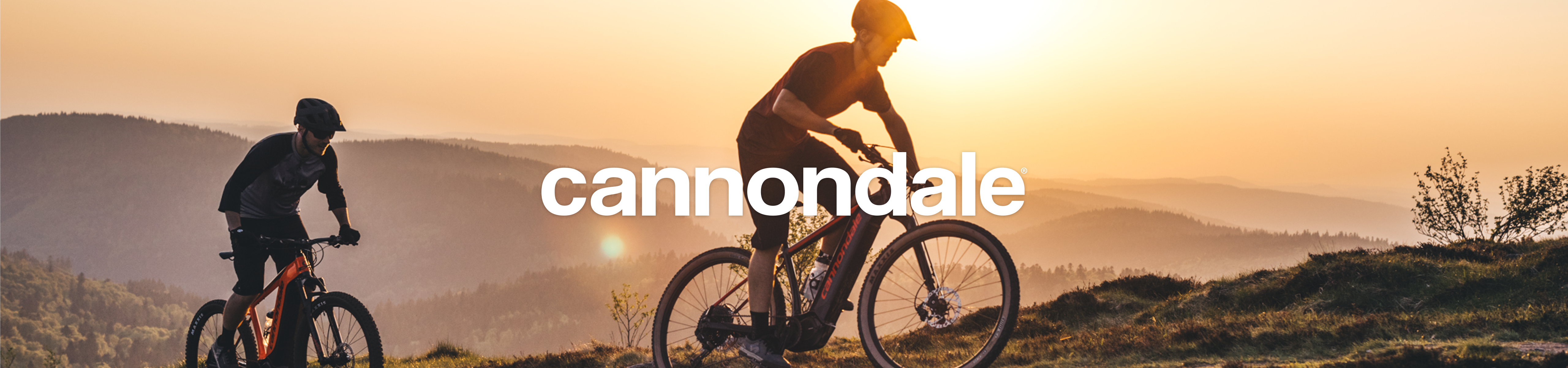 Zwei Mountainbiker fahren bei Sonnenuntergang über einen Hügel vor einer weiten Hügellandschaft. In der Bildmitte steht das weiße Logo von „cannondale“.